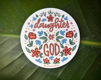 May include: A circular sticker featuring the text "daughter OF GOD" in a decorative font, surrounded by red, blue, and green floral designs. The sticker is white and rests on a green leaf.