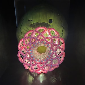 May include: A pink and white beaded sphere with a light inside, sitting in front of a green plush toy with a mustache.