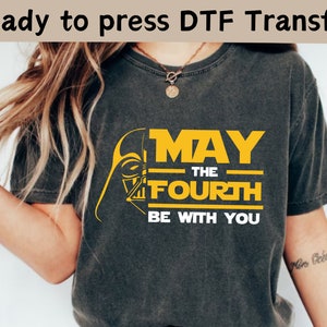 Puede incluir: Una camiseta gris oscura con un gráfico amarillo que dice "Que la fuerza te acompañe" y una silueta negra del casco de Darth Vader.