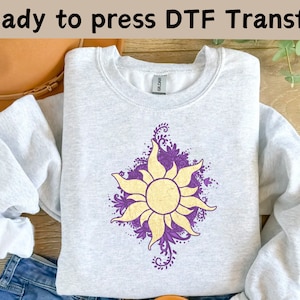 Könnte beinhalten: Ein graues Sweatshirt mit einem lila und goldenen Sonnenmotiv. Die Sonne hat ein florales Muster drumherum. Der Text "Ready to press DTF Transfer" befindet sich oben im Bild.