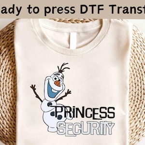 以下が含まれることがあります： クリーム色のTシャツ。雪だるまのオラフの漫画の画像と「PRINCESS SECURITY」の白黒の文字がプリントされています。「Ready to press DTF Transfer」というフレーズが上部に印刷されています。