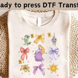 May include: Cream-colored t-shirt featuring a DTF transfer design. The design includes a cartoon princess in a purple dress, a green chameleon, bows, a tower, and a sun. The text "Ready to press DTF Transfer" is at the top.