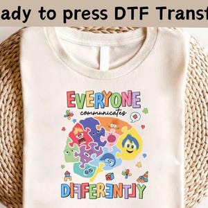 以下が含まれることがあります： 「Everyone Communicates Differently」とカラフルな文字で書かれたクリーム色のTシャツ。パズルのピースでできた脳と漫画のキャラクターがデザインされています。「Ready to press DTF Transfer」の文字が上部にあります。