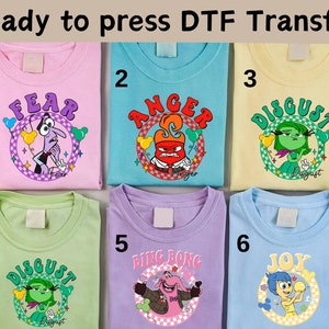 以下が含まれることがあります： 映画「インサイド・ヘッド」のキャラクターがプリントされた、明るい色のTシャツ6枚。キャラクターは、恐怖、怒り、嫌悪、喜び、ビング・ボンです。Tシャツは1から6まで番号が付けられています。