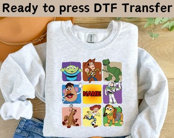 May include: A gray sweatshirt with a colorful graphic print of characters from the Toy Story movie franchise. The graphic includes Woody, Buzz Lightyear, Rex, Mr. Potato Head, Jessie, and Slinky Dog. The text "NAME" is printed in a yellow box.
