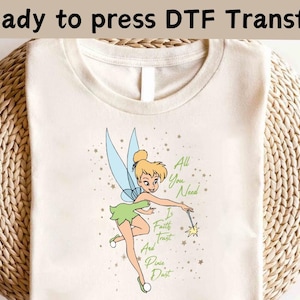 Könnte beinhalten: Ein weißes T-Shirt mit einem Transferdruck von Tinkerbell, einer Fee mit grünen Flügeln und einem grünen Kleid, die einen Zauberstab hält. Der Text "All You Need Is Faith, Trust, And Pixie Dust" ist auf dem Shirt gedruckt.
