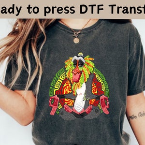 Może przedstawiać: Szary t-shirt z kolorowym nadrukiem graficznym przedstawiającym animowaną małpę medytującą. Małpa jest otoczona geometrycznymi wzorami w kolorze czerwonym, żółtym i zielonym. Tekst "Ready to press DTF Transfer" jest widoczny w górnej części obrazu.