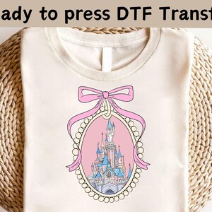 May include: A white t-shirt with a pink and white design of a castle with a pink bow on top. The design is in a frame with a pearl border.