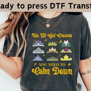 以下が含まれることがあります： 「We All Got Crowns You Need To Calm Down」というテキストと王冠のデザインが施された濃いグレーのTシャツです。