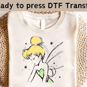 May include: Cream-colored t-shirt featuring a sketch of a fairy with yellow hair and a green dress. The words "Ready to press DTF Transfer" are printed at the top. The fairy is drawn in black ink with delicate lines and details.