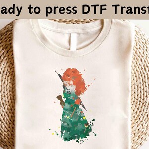 Puede incluir: Una camiseta blanca con una imagen al estilo acuarela de una mujer pelirroja con un vestido verde. El texto "Ready to press DTF Transfer" está en la parte superior de la imagen.