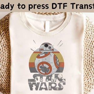 Puede incluir: Camiseta color crema con un gráfico de BB-8 en naranja y gris, sobre un diseño retro de puesta de sol. El logotipo de Star Wars está impreso debajo. El texto "Ready to press DTF Transfer" está en la parte superior.