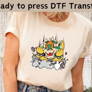 May include: A cream colored t-shirt with a cartoon image of Bowser from the Super Mario Bros. video game series. Bowser is depicted in a green and yellow costume with a red mouth and is shown breaking through a wall.