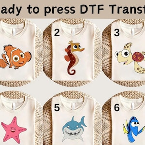 May include: Six white t-shirts with different cartoon images of sea creatures. The shirts are numbered 1 through 6. The images include a clownfish, a seahorse, a sea turtle, a starfish, a shark, and a blue tang.