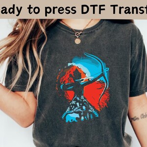 May include: Dark gray t-shirt featuring a graphic of an archer with a bow and arrow, set against a backdrop of red mountains and a blue sky. The design includes a bear and foliage. Text at the top reads "Ready to press DTF Transfer."