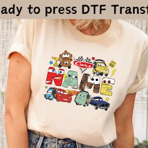 May include: A light beige t-shirt with a Cars themed design featuring the word "NAME" in large letters with various Cars characters surrounding the letters.