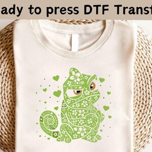 Könnte beinhalten: Ein weißes T-Shirt mit einem grünen Chamäleon-Design. Das Chamäleon besteht aus floralen Mustern und hat einen mürrischen Ausdruck. Das Design ist bereit, mit einem DTF-Transfer gepresst zu werden.