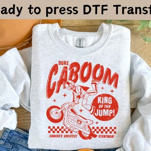May include: Gray sweatshirt with a red graphic print of a cartoon character performing a stunt on a motorcycle. The text on the graphic reads "Duke Caboom, King of the Jump! Canada's Greatest Stuntman!"