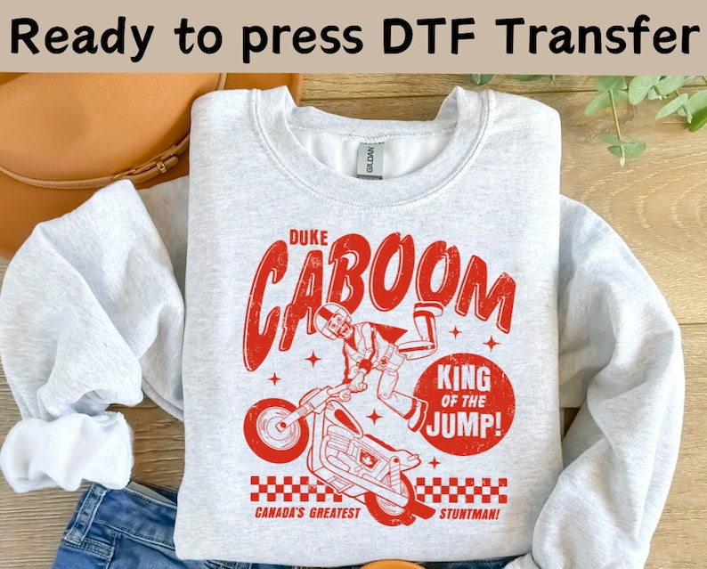 May include: Gray sweatshirt with a red graphic print of a cartoon character performing a stunt on a motorcycle. The text on the graphic reads "Duke Caboom, King of the Jump! Canada's Greatest Stuntman!"