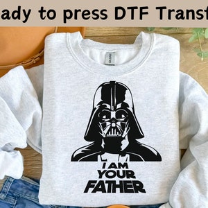 以下が含まれることがあります： ダース・ベイダーのヘルメットの黒いグラフィックと「I AM YOUR FATHER」の文字が入ったライトグレーのスウェットシャツ。「Ready to press DTF Transfer」の文字が上部にあります。スウェットシャツは折りたたまれており、ベージュの帽子とブルージーンズの一部が見えています。