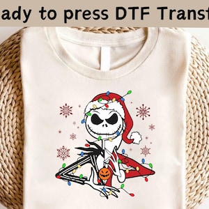 May include: A cream-coloured t-shirt with a DTF transfer design of Jack Skellington in a Santa hat, decorated with Christmas lights and snowflakes. The text "Ready to press DTF Transfer" is at the top. The design includes a pumpkin.