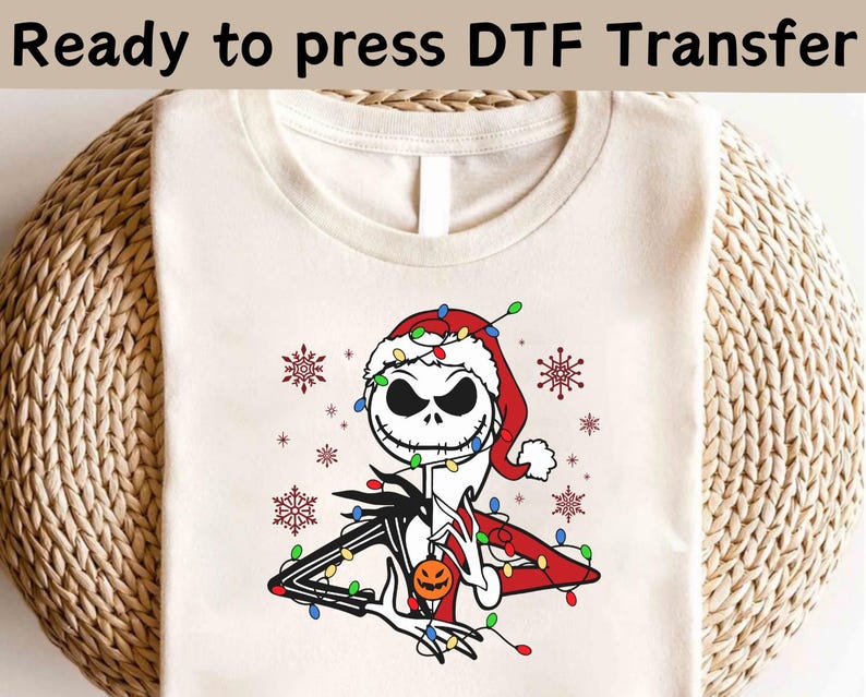 May include: A cream-coloured t-shirt with a DTF transfer design of Jack Skellington in a Santa hat, decorated with Christmas lights and snowflakes. The text "Ready to press DTF Transfer" is at the top. The design includes a pumpkin.