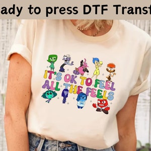以下が含まれることがあります： インサイド・ヘッドのキャラクターがカラフルに描かれた白いTシャツ。グラフィックには「It's OK to Feel All the Feels」というテキストが含まれています。