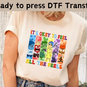 以下が含まれることがあります： インサイド・ヘッドのキャラクターがカラフルなグラフィックで描かれた白いTシャツ。グラフィックには「すべての感情を感じることは大丈夫」と書かれています。
