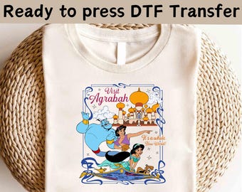 May include: Cream-colored t-shirt featuring a colorful graphic from the movie Aladdin. The graphic includes the text "Visit Agrabah" and "It's a whole New World!" along with illustrations of the Genie, Jasmine, and the city of Agrabah.