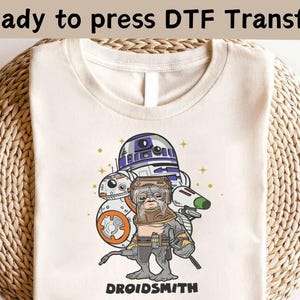 Op de afbeelding: Een wit T-shirt met een cartoonillustratie van Star Wars-personages R2-D2, C-3PO en een droid met de tekst "DROIDSMITH".