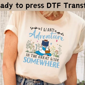 May include: A cream colored t-shirt with a blue and white graphic print of a girl reading a book in a field of flowers. The text on the shirt reads "I want adventure in the great wide somewhere."