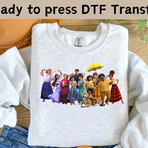 May include: A gray sweatshirt with a print of the characters from the animated film "Encanto". The characters are standing in front of a yellow umbrella. The text "Ready to press DTF Transfer" is visible at the top of the image.