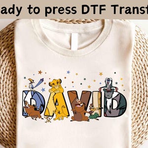 Może przedstawiać: Kremowa koszulka z napisem "DAVID" w kolorowej, kreskówkowej czcionce. Litery ozdobione są postaciami z popularnej animacji, z gwiazdami rozrzuconymi wokół imienia. U góry znajduje się napis "Ready to press DTF Transfer".