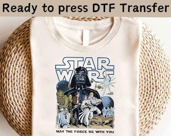 May include: Cream-coloured t-shirt with a Star Wars graphic. The design features Darth Vader, other characters, and the text "STAR WARS" in blue. The phrase "MAY THE FORCE BE WITH YOU" is printed below the graphic.