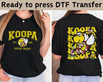 May include: Black t-shirt featuring a yellow and white graphic of a Koopa Troopa from Super Mario Bros. The graphic reads "Koopa Est. 1985 Super Mario". The back of the shirt has a repeating pattern of the Koopa Troopa graphic with stars, mushrooms, and question blocks.