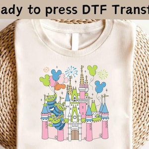 May include: Cream-colored t-shirt featuring a colorful graphic of a fairytale castle with green, pink, and blue accents. The design includes cartoon aliens holding balloons and fireworks. Text at the top reads "Ready to press DTF Transfer."