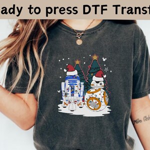 May include: Dark gray t-shirt featuring a festive graphic with R2-D2 and BB-8 wearing Santa hats, standing near a Christmas tree. The text "Ready to press DTF Transfer" is at the top. The design is perfect for holiday apparel.