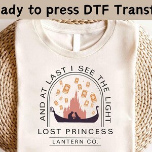 Könnte beinhalten: Ein beiges T-Shirt mit einem schwarz-weißen Design, das eine Silhouette eines Paares in einem Boot mit umherfliegenden Laternen zeigt. Der Text "And at last I see the light" ist in einem Kreis um das Paar geschrieben. Unter dem Design steht der Text "Lost Princess Lantern Co." in Schwarz.