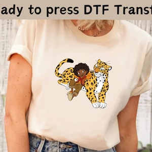 May include: A cream-colored t-shirt with a cartoon illustration of a young person riding on a jaguar. The jaguar is yellow and black with black spots. The young person is wearing a brown shirt and has brown hair. The text "Ready to press DTF Transfer" is at the top of the image.