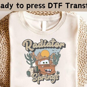 May include: A beige t-shirt with a graphic design of a cartoon car in a desert landscape. The text "Radiator Springs" is printed below the car.