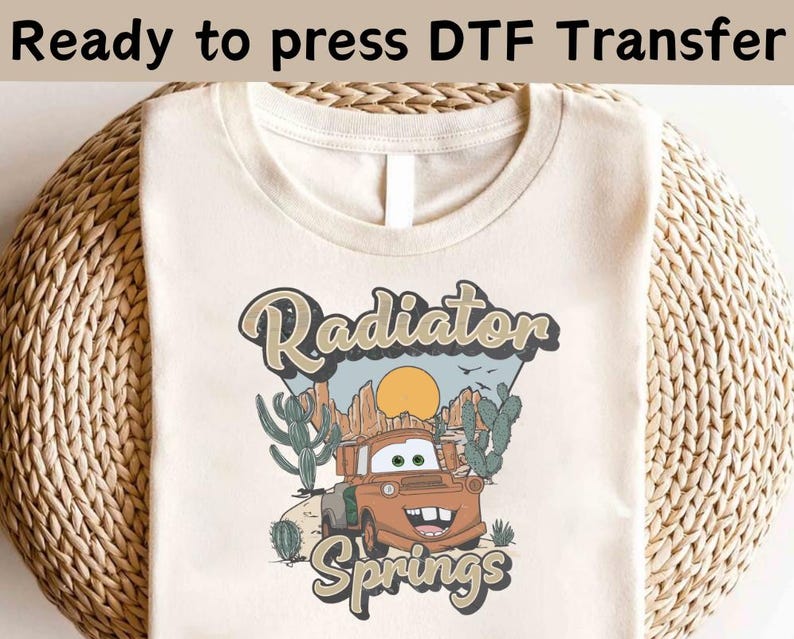 May include: A beige t-shirt with a graphic design of a cartoon car in a desert landscape. The text "Radiator Springs" is printed below the car.