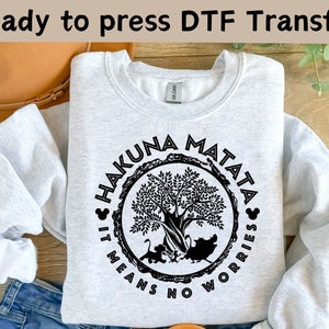 Op de afbeelding: Een grijs sweatshirt met een zwarte print van een boom met de woorden "Hakuna Matata" eromheen. De tekst "It means no worries" staat ook op het sweatshirt gedrukt.