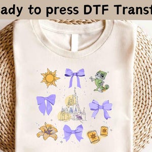 May include: A beige t-shirt with a purple bow, a green chameleon, a yellow sun, a yellow flower, a castle, and a purple bow. The text "Ready to press DTF Transfer" is at the top of the image.