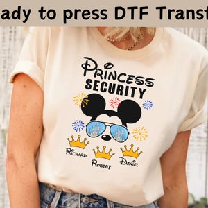 Może przedstawiać: Biała koszulka z nadrukiem Mickey Mouse'a i tekstem "Princess Security". Mickey Mouse nosi okulary przeciwsłoneczne z odbiciem zamku Kopciuszka. Pod spodem znajdują się trzy złote korony z nazwami "Richard", "Robert" i "Daniel".