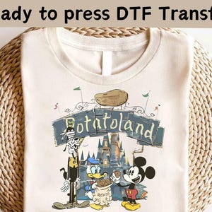 May include: A beige t-shirt with a cartoon graphic of Mickey Mouse, Goofy, and Donald Duck. The graphic features a castle and the text "PotatoLand".