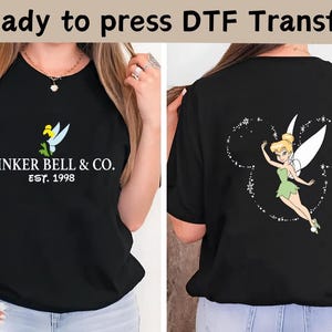 Könnte beinhalten: Schwarzes T-Shirt mit einem Tinker Bell-Motiv auf der Vorder- und Rückseite. Auf der Vorderseite steht der Text "TINKER BELL & CO. EST. 1998" mit einem Tinker Bell-Design. Auf der Rückseite ist Tinker Bell in einer Mickey Mouse-Umrisslinie zu sehen.