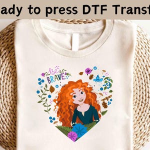 以下が含まれることがあります： クリーム色のTシャツ。赤い巻き毛の女の子の漫画イラストがハート型の花柄の中に描かれています。「Be Brave」の文字が女の子の頭の上にあります。「Ready to press DTF Transfer」の文字が上部にあります。