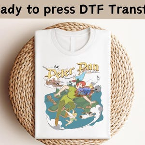 May include: A white t-shirt featuring a colorful graphic print of Peter Pan, Tinker Bell, and other characters from the story. The text Peter Pan is displayed in a stylized yellow font above the characters, with a blue sea and green land below.