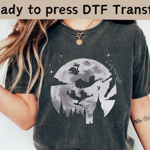 Może przedstawiać: Szary t-shirt z białym nadrukiem przedstawiającym sylwetki Piotrusia Pana, Dzwoneczka i Kapitana Haka lecących przed pełnią księżyca. Nadruk zawiera również sylwetkę panoramy miasta. Tekst "Ready to press DTF Transfer" znajduje się w górnej części obrazu.