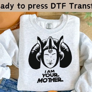 以下が含まれることがあります： スタイリッシュな女性の顔の黒いグラフィックと「I AM YOUR MOTHER」のテキストが特徴のライトグレーのスウェットシャツ。スウェットシャツは、すぐにプレスできるDTF転写です。